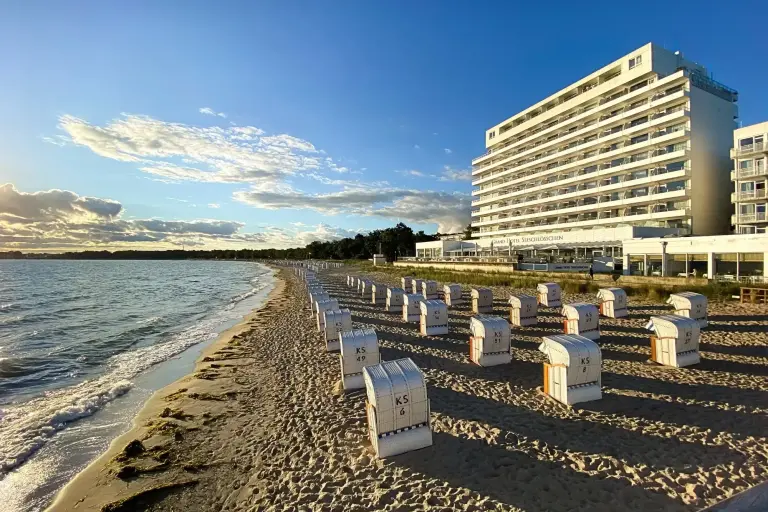 Ein modernes Hotel direkt am Strand der Ostsee, im Vordergrund viele Strandkörbe in der Abendsonne.